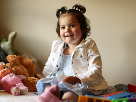 A toddler girl sits on her toys and smiles.