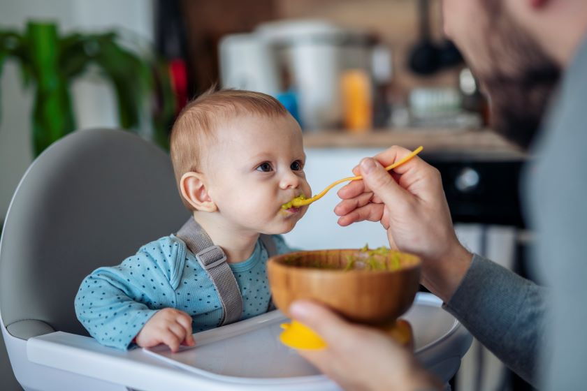 A baby is sitting in a high chair while his dad feeds him.