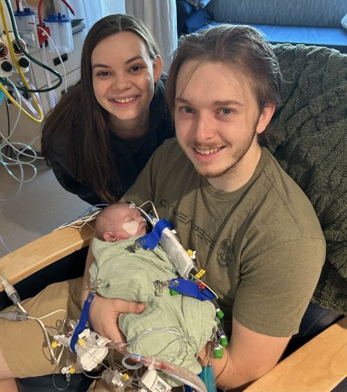 David's dad holds him in a hospital rocking chair. David's mom poses alongside them.